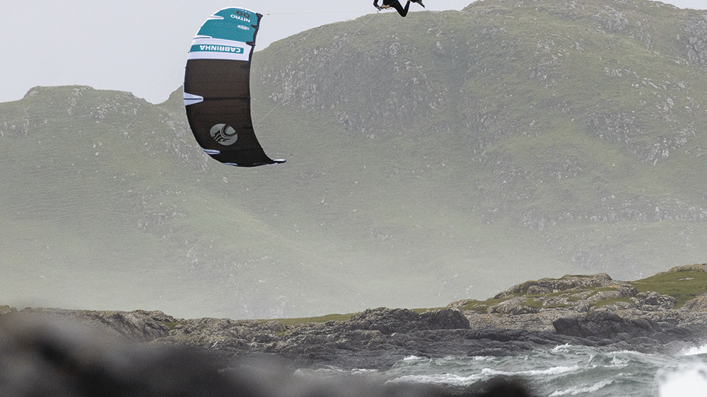 Directional vs Twin Tip Kiteboards…Which Should You Ride? Kiteboarder jumping over waves with scenic mountains in background.
