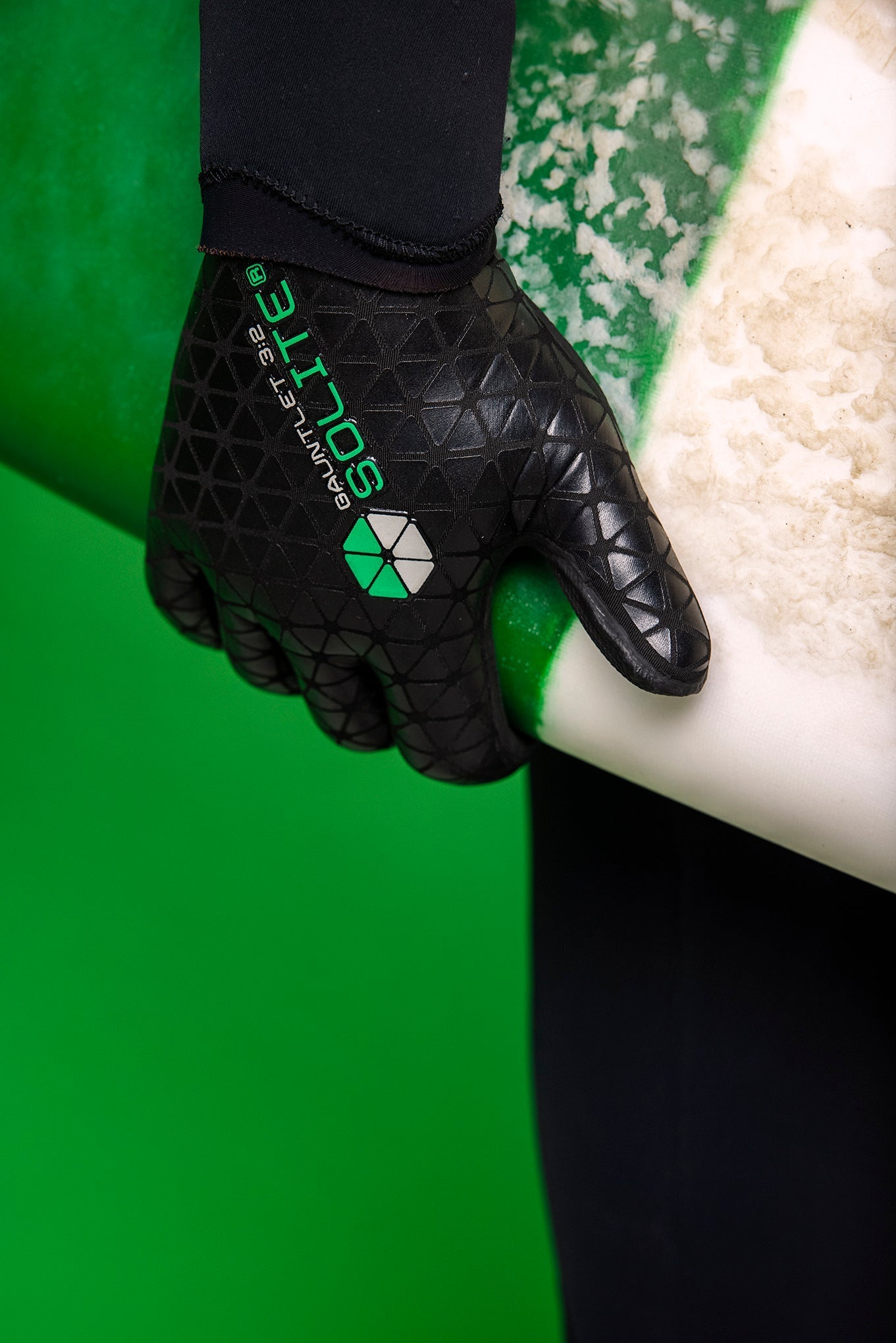 A hand wearing a 3:2 Gauntlet Wetsuit Glove, holding a surfboard with vibrant green background.