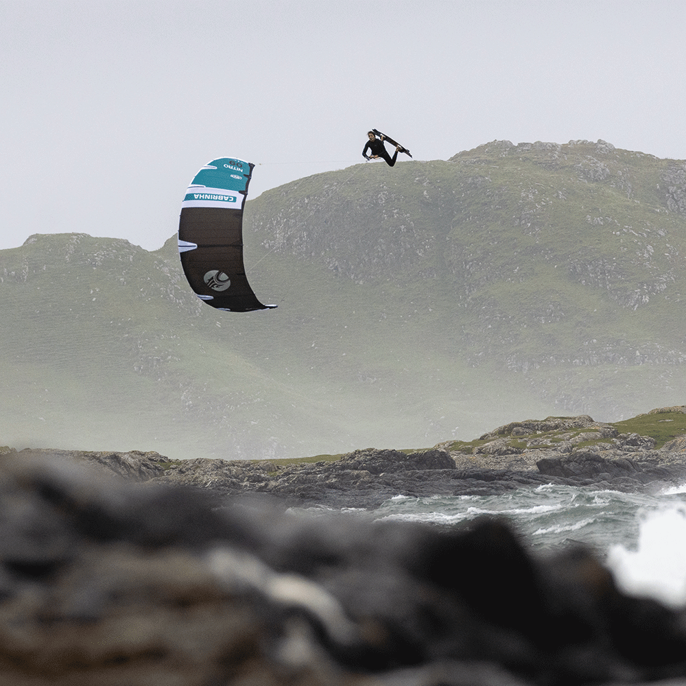 Directional vs Twin Tip Kiteboards…Which Should You Ride? Kiteboarder jumping over waves with scenic mountains in background.
