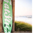 Fatstick AirStick 10'6 Inflatable SUP Package 2026 leaning against wooden wall by the beach.